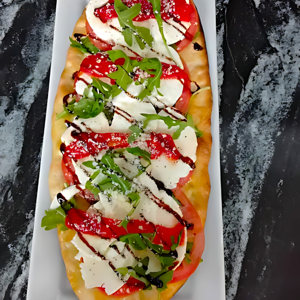 A long flatbread pizza with mozzarella, tomatoes, arugula, and balsamic glaze on a white platter.